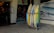 A collection of surfboards is neatly arranged in a rack outside a surf shop. The shop has a festive holiday greeting sign and posters advertising surfing events. The entrance to the shop is visible, showing a warmly lit interior with various surfing merchandise displayed. A poster on the wall features a wave with dates and text relating to a surfing competition.