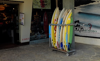 A collection of surfboards is neatly arranged in a rack outside a surf shop. The shop has a festive holiday greeting sign and posters advertising surfing events. The entrance to the shop is visible, showing a warmly lit interior with various surfing merchandise displayed. A poster on the wall features a wave with dates and text relating to a surfing competition.