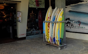 A collection of surfboards is neatly arranged in a rack outside a surf shop. The shop has a festive holiday greeting sign and posters advertising surfing events. The entrance to the shop is visible, showing a warmly lit interior with various surfing merchandise displayed. A poster on the wall features a wave with dates and text relating to a surfing competition.