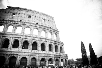 A black-and-white image featuring the Colosseum in Rome, showcasing its ancient arched architecture. A crowd of people is gathered in front of the monument. There are tall trees visible on the right side, adding contrast to the stone structure.
