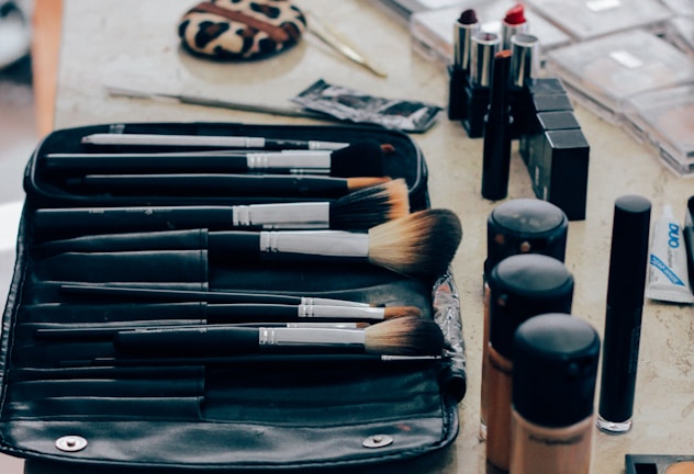 A collection of makeup tools and cosmetics organized on a table. Multiple makeup brushes of varying sizes are placed neatly in a black brush holder. Lipsticks with red and neutral shades are standing upright beside several bottles of liquid foundation in different tones. A leopard-print compact and several other makeup items, such as mascara and concealer, can also be seen.