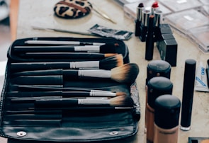 A collection of makeup tools and cosmetics organized on a table. Multiple makeup brushes of varying sizes are placed neatly in a black brush holder. Lipsticks with red and neutral shades are standing upright beside several bottles of liquid foundation in different tones. A leopard-print compact and several other makeup items, such as mascara and concealer, can also be seen.
