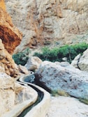 A rocky desert landscape with a narrow irrigation channel winding through. Large boulders and a background of rugged, steep cliffs dominate the scene. Lush green palm trees are visible in the distance, providing a stark contrast to the arid environment.