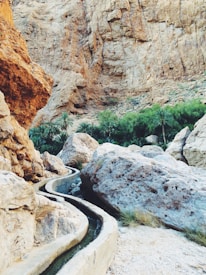 A rocky desert landscape with a narrow irrigation channel winding through. Large boulders and a background of rugged, steep cliffs dominate the scene. Lush green palm trees are visible in the distance, providing a stark contrast to the arid environment.
