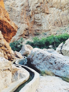A rocky desert landscape with a narrow irrigation channel winding through. Large boulders and a background of rugged, steep cliffs dominate the scene. Lush green palm trees are visible in the distance, providing a stark contrast to the arid environment.
