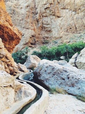 A rocky desert landscape with a narrow irrigation channel winding through. Large boulders and a background of rugged, steep cliffs dominate the scene. Lush green palm trees are visible in the distance, providing a stark contrast to the arid environment.