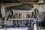 A rustic workshop wall displaying an assortment of vintage woodworking tools. Numerous chisels, saws, and hand drills are organized neatly, with a large hand saw prominently hanging above the rest. A coil of rope is also visible, adding to the aged, well-used feel of the workspace.