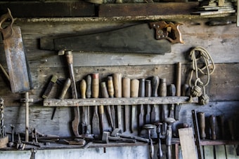 A rustic workshop wall displaying an assortment of vintage woodworking tools. Numerous chisels, saws, and hand drills are organized neatly, with a large hand saw prominently hanging above the rest. A coil of rope is also visible, adding to the aged, well-used feel of the workspace.