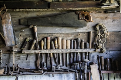 A rustic workshop wall displaying an assortment of vintage woodworking tools. Numerous chisels, saws, and hand drills are organized neatly, with a large hand saw prominently hanging above the rest. A coil of rope is also visible, adding to the aged, well-used feel of the workspace.