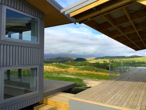 A modern architectural structure with large glass windows and corrugated metal siding is situated in a rural setting. A spacious wooden deck extends outward, overlooking rolling hills and pastures under a partly cloudy sky.