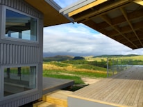 A modern architectural structure with large glass windows and corrugated metal siding is situated in a rural setting. A spacious wooden deck extends outward, overlooking rolling hills and pastures under a partly cloudy sky.
