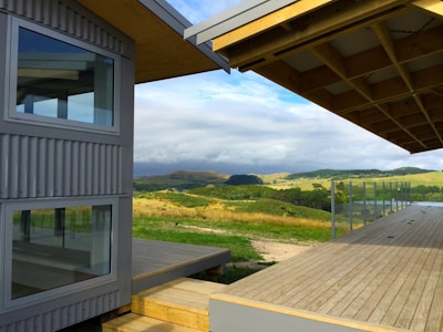 A modern architectural structure with large glass windows and corrugated metal siding is situated in a rural setting. A spacious wooden deck extends outward, overlooking rolling hills and pastures under a partly cloudy sky.