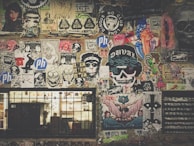 A vibrant collage of street art and posters covers a wall, featuring a mix of colorful designs, graffiti, and illustrations. Icons include skulls, cartoonish faces, animals, and various logos. The artwork is layered, with some stickers and hand-drawn images overlapping others, creating a chaotic yet artistic arrangement. Below the art, a barred window with graffiti adds to the urban aesthetic.