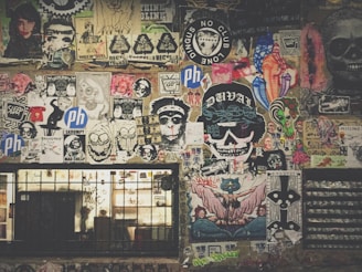 A vibrant collage of street art and posters covers a wall, featuring a mix of colorful designs, graffiti, and illustrations. Icons include skulls, cartoonish faces, animals, and various logos. The artwork is layered, with some stickers and hand-drawn images overlapping others, creating a chaotic yet artistic arrangement. Below the art, a barred window with graffiti adds to the urban aesthetic.