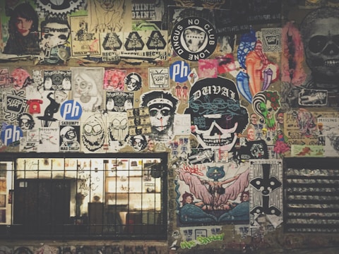 A vibrant collage of street art and posters covers a wall, featuring a mix of colorful designs, graffiti, and illustrations. Icons include skulls, cartoonish faces, animals, and various logos. The artwork is layered, with some stickers and hand-drawn images overlapping others, creating a chaotic yet artistic arrangement. Below the art, a barred window with graffiti adds to the urban aesthetic.