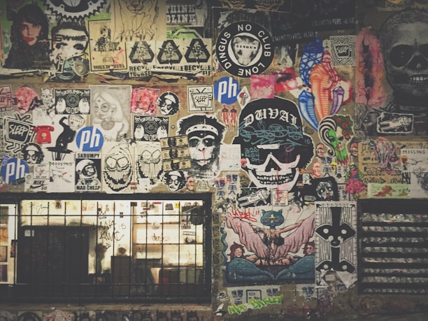 A vibrant collage of street art and posters covers a wall, featuring a mix of colorful designs, graffiti, and illustrations. Icons include skulls, cartoonish faces, animals, and various logos. The artwork is layered, with some stickers and hand-drawn images overlapping others, creating a chaotic yet artistic arrangement. Below the art, a barred window with graffiti adds to the urban aesthetic.