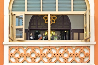 A window with two open shutters revealing an interior space adorned with intricate patterns. A geometric design dominates the lower part of the frame, while a person in traditional attire is seen through the window. The interior is softly lit with a warm glow, and a decorative, arched design forms part of the backdrop.