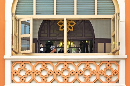 A window with two open shutters revealing an interior space adorned with intricate patterns. A geometric design dominates the lower part of the frame, while a person in traditional attire is seen through the window. The interior is softly lit with a warm glow, and a decorative, arched design forms part of the backdrop.