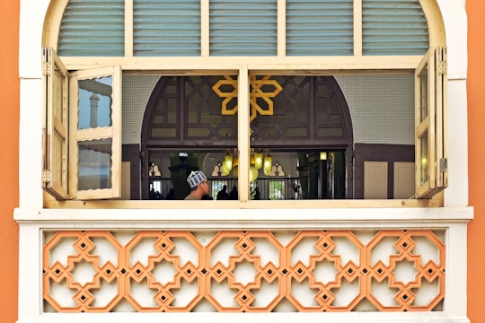 A window with two open shutters revealing an interior space adorned with intricate patterns. A geometric design dominates the lower part of the frame, while a person in traditional attire is seen through the window. The interior is softly lit with a warm glow, and a decorative, arched design forms part of the backdrop.