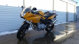 A yellow motorcycle is parked on a wet concrete surface next to a row of outdoor storage units. The motorcycle features a sleek design with black accents and a visible engine. The surrounding area is partially covered with snow, and the storage unit doors are metallic and shutter-like.