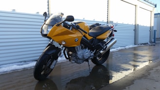 A yellow motorcycle is parked on a wet concrete surface next to a row of outdoor storage units. The motorcycle features a sleek design with black accents and a visible engine. The surrounding area is partially covered with snow, and the storage unit doors are metallic and shutter-like.