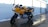 A yellow motorcycle is parked on a wet concrete surface next to a row of outdoor storage units. The motorcycle features a sleek design with black accents and a visible engine. The surrounding area is partially covered with snow, and the storage unit doors are metallic and shutter-like.