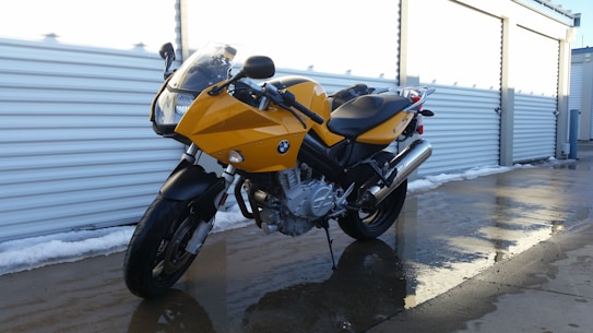 A yellow motorcycle is parked on a wet concrete surface next to a row of outdoor storage units. The motorcycle features a sleek design with black accents and a visible engine. The surrounding area is partially covered with snow, and the storage unit doors are metallic and shutter-like.