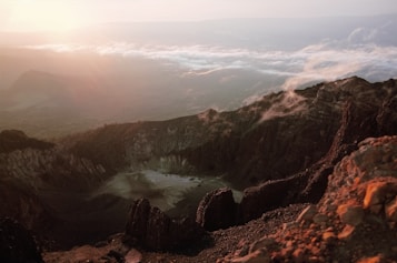 A breathtaking view of a volcanic crater surrounded by rocky terrain. The sunrise casts warm light over the scene, illuminating the rugged landscape with shades of orange and pink. Mist and clouds hover near the peaks, adding a mystical quality to the atmosphere.