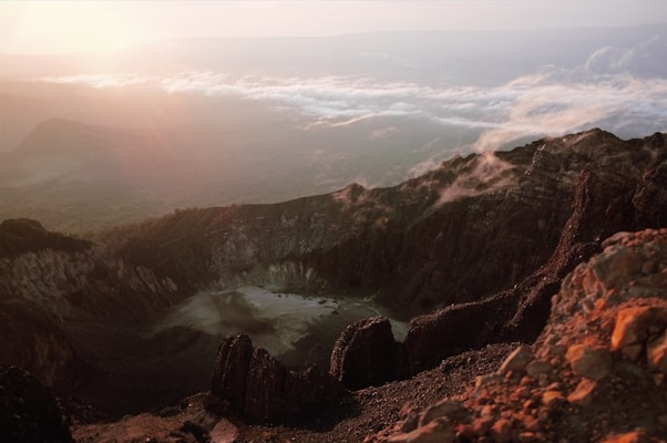 A breathtaking view of a volcanic crater surrounded by rocky terrain. The sunrise casts warm light over the scene, illuminating the rugged landscape with shades of orange and pink. Mist and clouds hover near the peaks, adding a mystical quality to the atmosphere.
