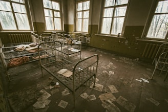 A dimly lit, abandoned room with several rusty metal bed frames scattered throughout. The walls are peeling, and the floor is covered with scattered papers and debris. The windows are dirty and allow bleak light to filter in, casting shadows across the room.