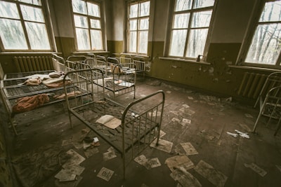 A dimly lit, abandoned room with several rusty metal bed frames scattered throughout. The walls are peeling, and the floor is covered with scattered papers and debris. The windows are dirty and allow bleak light to filter in, casting shadows across the room.