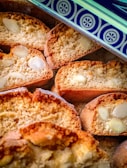 Several slices of almond biscotti are displayed, showing a crunchy texture with visible almond pieces. The top edge of a decorative tin with a blue and white pattern is partially visible.