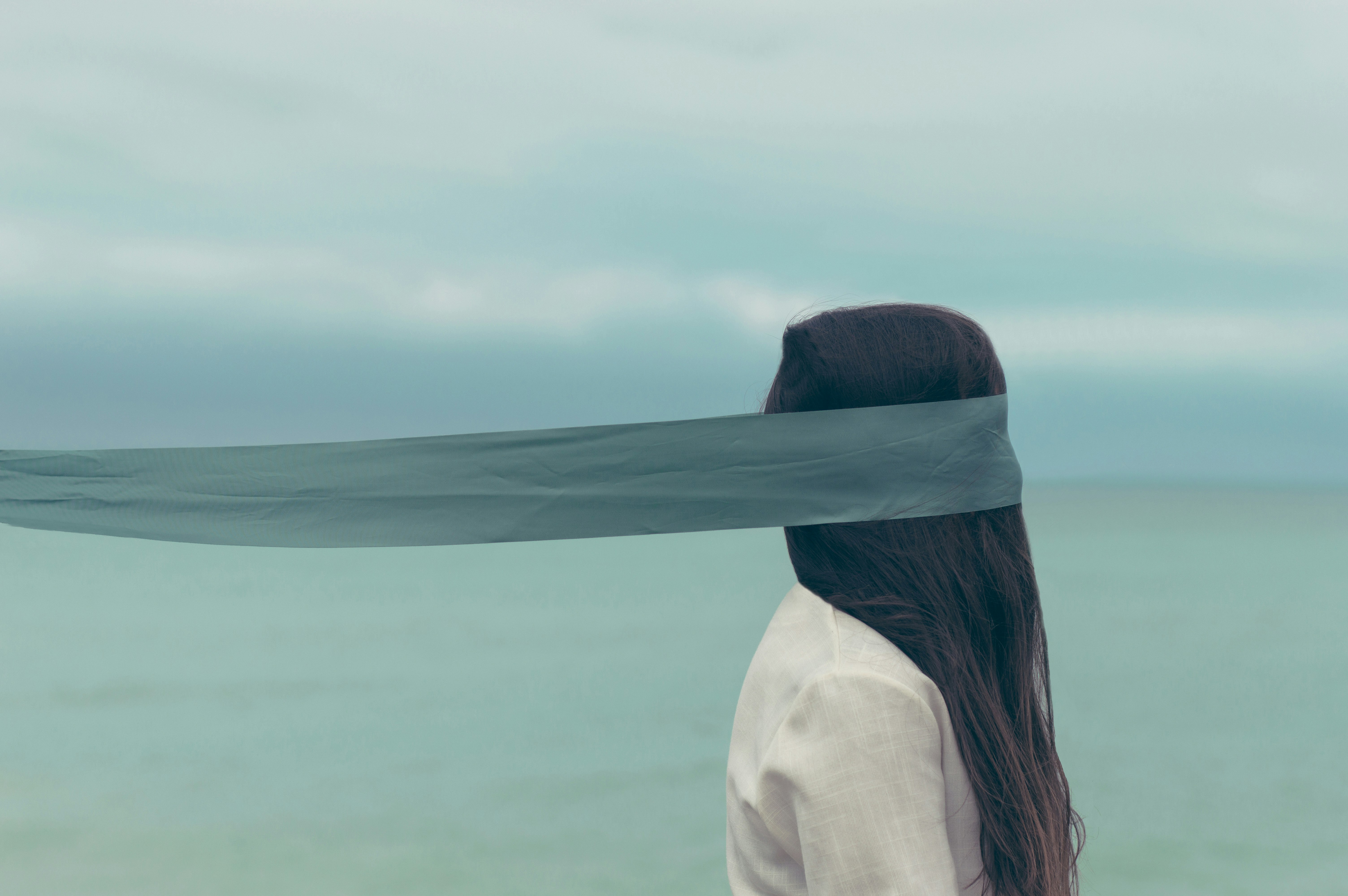 An image of a person with long brown hair and a white shirt. Around this person's head is a blue scarf. The arrangement of scarf and hair obscure the person's face, and it is unclear which direction they are facing. Behind is an open sky and expanse of water.