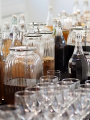 A display of various glass bottles and jars filled with different liquids, some opaque and others transparent. In the foreground, there are several rows of empty clear glass cups.