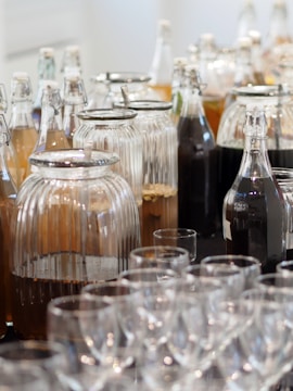 A display of various glass bottles and jars filled with different liquids, some opaque and others transparent. In the foreground, there are several rows of empty clear glass cups.
