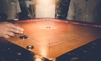 A game of carrom is being played on a wooden board with several carrom men and a striker. One player's hand is visible, poised to flick the striker towards the carrom pieces. The setting appears cozy and dimly lit, suggesting an indoor environment with warm lighting. People are partially visible around the board, implying a casual gathering.