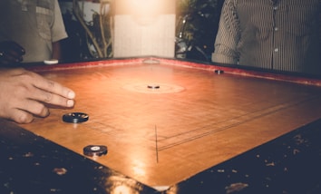 A game of carrom is being played on a wooden board with several carrom men and a striker. One player's hand is visible, poised to flick the striker towards the carrom pieces. The setting appears cozy and dimly lit, suggesting an indoor environment with warm lighting. People are partially visible around the board, implying a casual gathering.