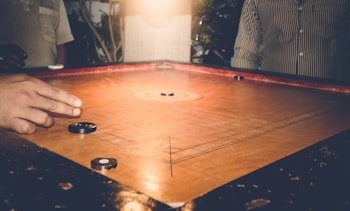 A game of carrom is being played on a wooden board with several carrom men and a striker. One player's hand is visible, poised to flick the striker towards the carrom pieces. The setting appears cozy and dimly lit, suggesting an indoor environment with warm lighting. People are partially visible around the board, implying a casual gathering.