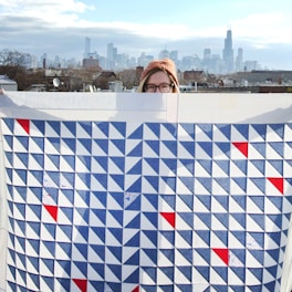 A person partially obscured by a large quilt with a geometric pattern. The quilt features blue, white, and red triangles arranged in a repetitive design. The person is wearing a knitted hat and glasses. Behind them is a cityscape with a skyline featuring tall buildings under a partly cloudy sky.