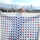 A person partially obscured by a large quilt with a geometric pattern. The quilt features blue, white, and red triangles arranged in a repetitive design. The person is wearing a knitted hat and glasses. Behind them is a cityscape with a skyline featuring tall buildings under a partly cloudy sky.