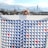 A person partially obscured by a large quilt with a geometric pattern. The quilt features blue, white, and red triangles arranged in a repetitive design. The person is wearing a knitted hat and glasses. Behind them is a cityscape with a skyline featuring tall buildings under a partly cloudy sky.