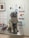 A pug sitting on a metallic examination table in a veterinary clinic, surrounded by posters and informational flyers about pet health and medications on the wall. The pug is wearing a black harness and has a slightly open mouth, giving an impression of a happy expression.