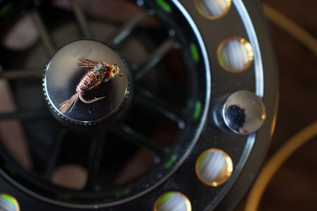 A close-up of a fly fishing reel with a detailed fly lure placed on top. The reel exhibits a mix of metallic and dark shades, with visible mechanical parts and a textured surface. The fly lure is intricately crafted, featuring delicate fibers and a metallic shine.