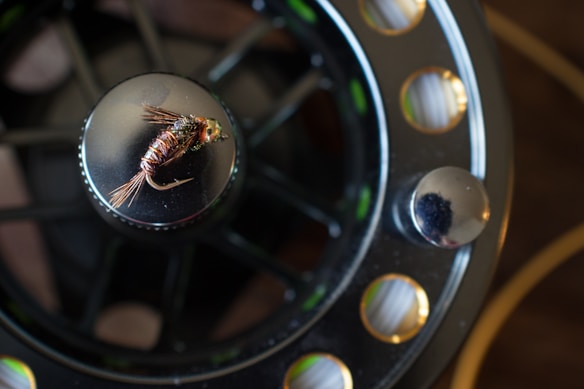 A close-up of a fly fishing reel with a detailed fly lure placed on top. The reel exhibits a mix of metallic and dark shades, with visible mechanical parts and a textured surface. The fly lure is intricately crafted, featuring delicate fibers and a metallic shine.