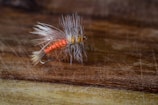A detailed view of a fishing fly with bright orange and yellow threads, complemented by feather-like bristles, all attached to a metal fish hook. The object is placed on a textured wooden surface that adds a rustic feel.