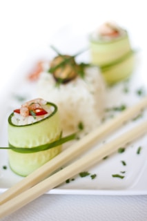 A culinary presentation featuring sushi rolls wrapped in thin cucumber slices with visible rice, shrimp, and garnished with herbs. The rolls are arranged on a white rectangular plate alongside a pair of wooden chopsticks. The background contains a blurred image of additional sushi rolls and sprinkles of green herbs.