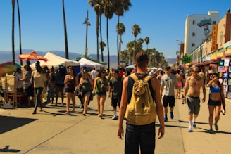 A busy boardwalk scene with a crowd of people walking in different directions. A man in the foreground wears a backpack and faces away from the camera. Various stands and shops line the street, with umbrellas, signs for pizza, and other attractions visible. Tall palm trees border the street on both sides under a clear blue sky.