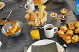 A table is set with an array of breakfast items, including croissants in a metal bowl, sliced baguette in a basket, glasses of orange juice, and a cup of coffee. Three lit candles add warmth to the setting. There are also cheese-topped savory pastries on a black plate.