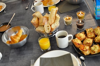 A table is set with an array of breakfast items, including croissants in a metal bowl, sliced baguette in a basket, glasses of orange juice, and a cup of coffee. Three lit candles add warmth to the setting. There are also cheese-topped savory pastries on a black plate.