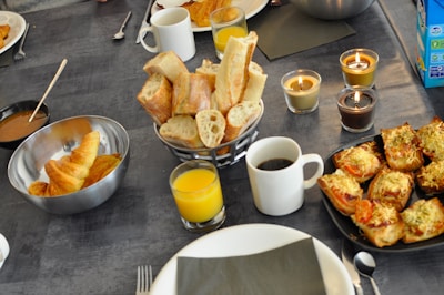 A table is set with an array of breakfast items, including croissants in a metal bowl, sliced baguette in a basket, glasses of orange juice, and a cup of coffee. Three lit candles add warmth to the setting. There are also cheese-topped savory pastries on a black plate.